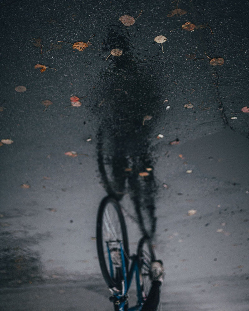 Nasser Asphalt mit Herbstlaub, darin spiegelt sich ein Radfahrer.