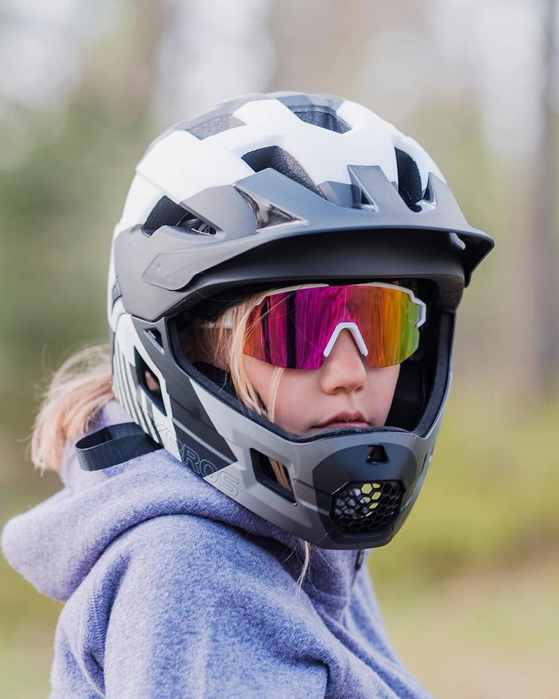Ein Kind trägt einen Fullface-Helm und eine verspiegelte Sportbrille, dahinter unscharfer Wald.